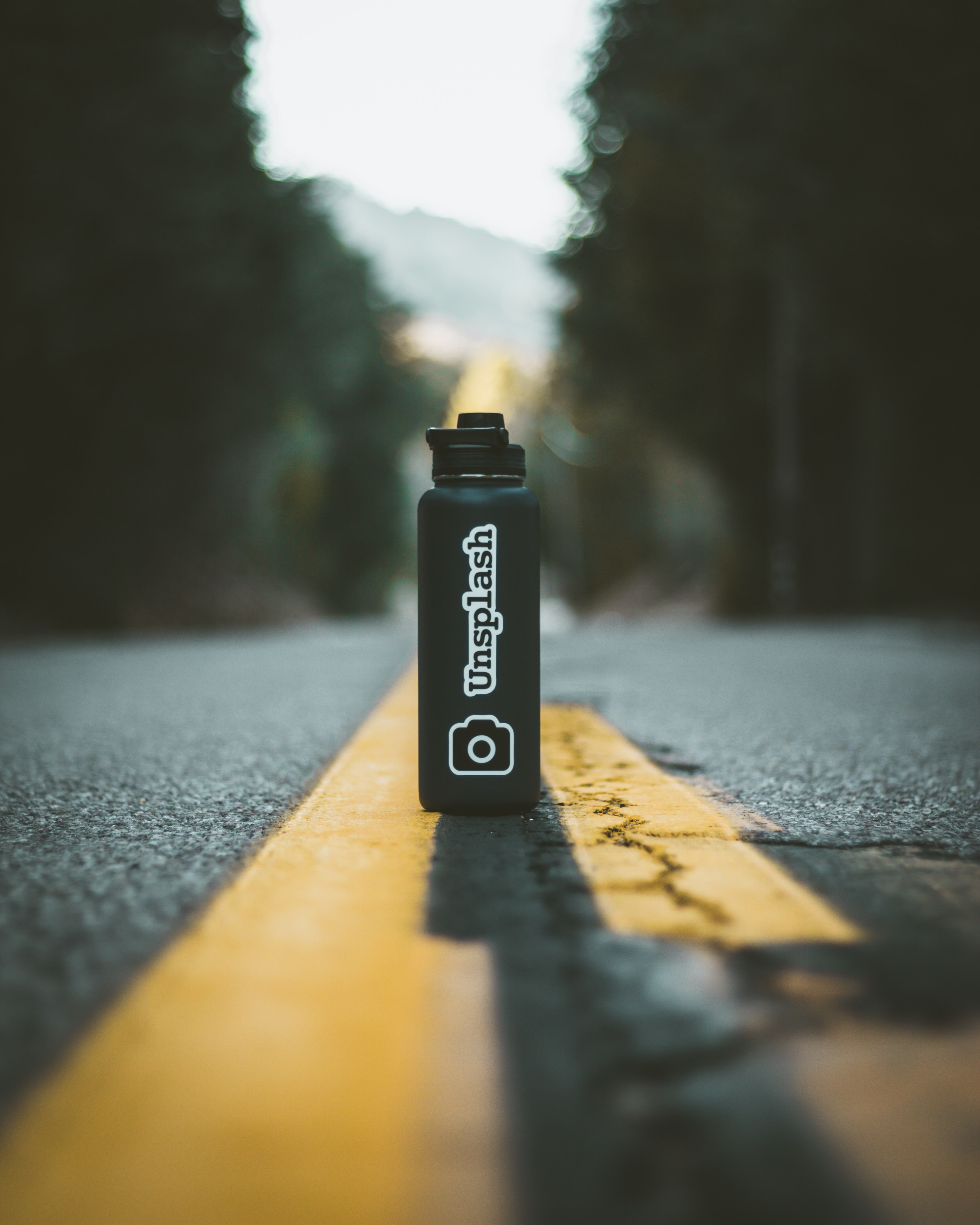 Photograph of a black metal water bottle sitting in the middle of a road. The water bottle has a sticker that says 'unsplash' above a sticker of a camera.